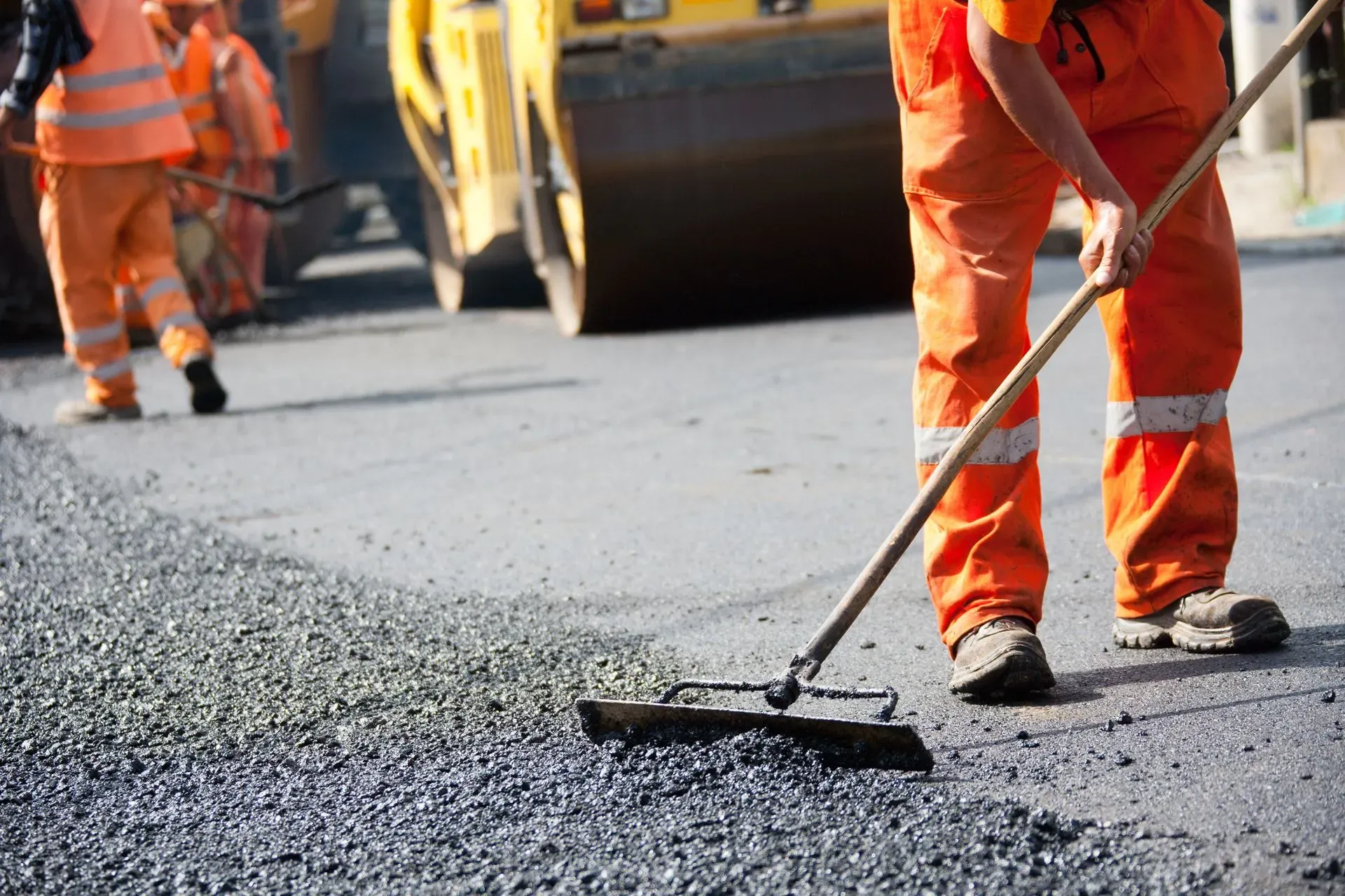 Paving crew member raking fresh hot asphalt by hand while a roller and crew work behind